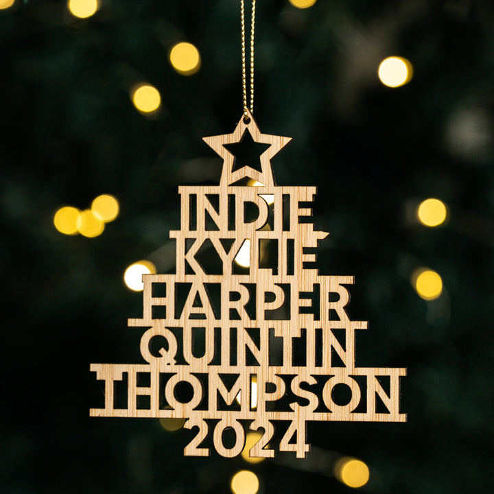 A custom-made wooden Christmas tree bauble with family names and the year 2024 inscribed, hanging from a tree with blurred lights in the background.