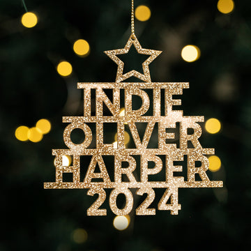 A gold, glittery Whānau Christmas Tree Bauble personalised with "INDIE OLIVER HARPER 2024" and topped with a star hangs against blurred yellow holiday lights.