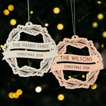 Two Yuletide Wreath Christmas Baubles hang on the tree; one reads "The Harris Family Christmas 2024," the other "The Wilsons Christmas 2024," with blurred festive lights twinkling in the background.