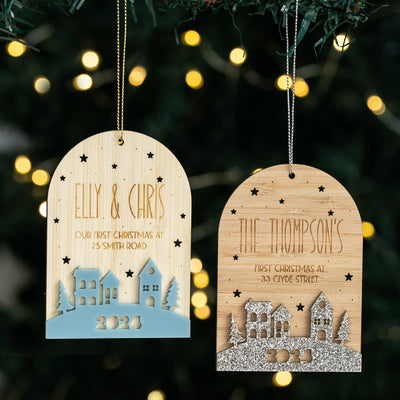 Two "Our First Home Christmas Globe" ornaments hang on a tree, each engraved with names, address, and 2024. Festive designs of houses and trees glow against blurred holiday lights.