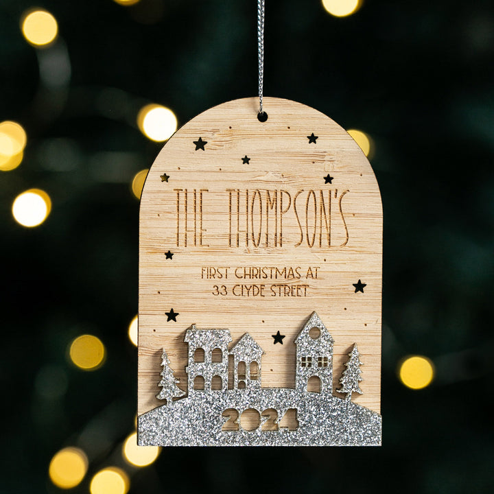 The Our First Home Christmas Globe ornament features "The Thompsons, First Christmas at 33 Clyde Street" with stars, houses, and "2021" in glitter, displayed against blurred festive lights.