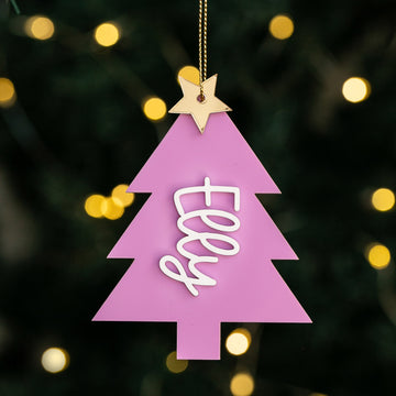 The Carnival Christmas Tree Bauble, a pink acrylic ornament shaped like a tree with a gold star and "Elly" in white cursive, hangs against softly glowing holiday lights.