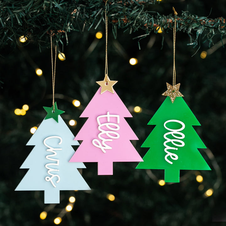 Three Christmas tree-shaped baubles in blue, pink, and green, each with a sparkling star on top and a custom name in script on the pink and green ones.