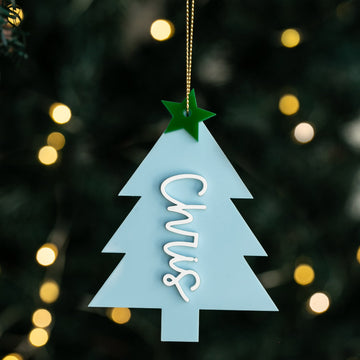 The Carnival Christmas Tree Bauble features a light blue design with a green star on top and "Chris" in white script, hanging from a gold string against blurred holiday lights in the background.