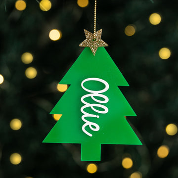 The Carnival Christmas Tree Bauble is a green acrylic ornament featuring "Ollie" in white script and a glittery gold star on top, shown hanging with blurred yellow lights in the background.