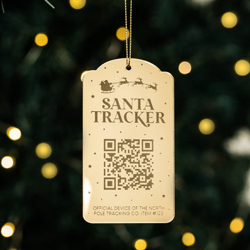 The Santa Tracker Christmas Bauble, with a gold finish, QR code, festive text, and a silhouette of Santa’s sleigh and reindeer, hangs on a Christmas tree adorned with blurred lights in the background.