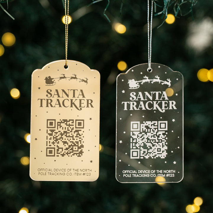 Two ornament tags—a Santa Tracker Christmas Bauble and a clear tag—hang on a Christmas tree. Each interactive bauble features a QR code, festive icons, and hanging string, with twinkling tree lights blurred in the background.