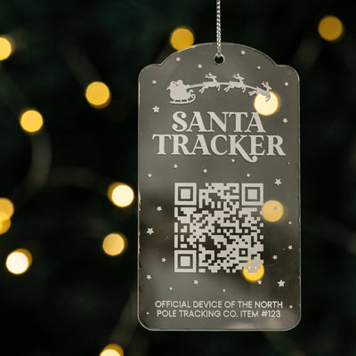 A Christmas bauble with a QR code and 'Santa Tracker' text, hanging from a string, with a blurred Christmas tree and lights in the background.