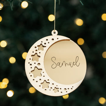 The Sweet Dreams Christmas Bauble is a crescent moon with star cutouts and the name Samuel at its center, hanging on a string. Blurred yellow lights twinkle in the background, making it an ideal first Christmas keepsake.