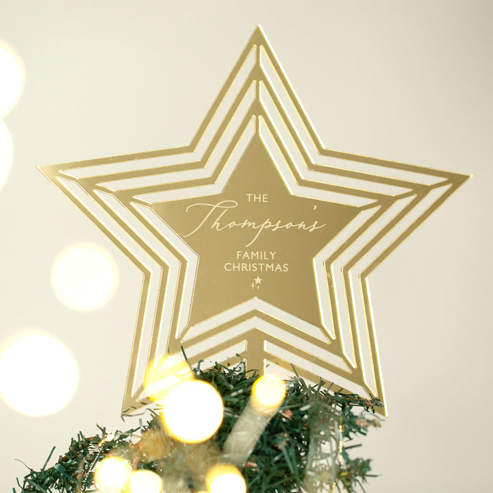 A gold Personalised Christmas Tree Topper in a star shape, engraved with "The Thompson’s Family Christmas," crowns a green tree adorned with soft yellow lights, adding a festive glow.