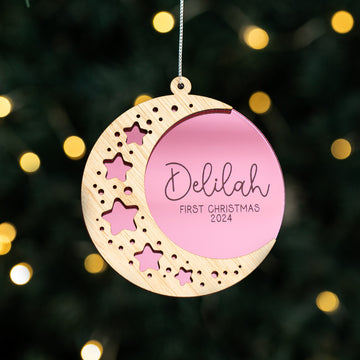 The Sweet Dreams Christmas Bauble is a bamboo crescent moon ornament with star cutouts and a pink center, personalised with "Delilah" and "First Christmas 2024," shown hanging before blurred tree lights.