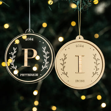Two Monogram Christmas Baubles hang on the tree. Each bauble features a name, initial, 2024, and leafy borders—a personalised ornament and charming keepsake from The Monogram Christmas Bauble collection. Blurred lights glow softly behind.