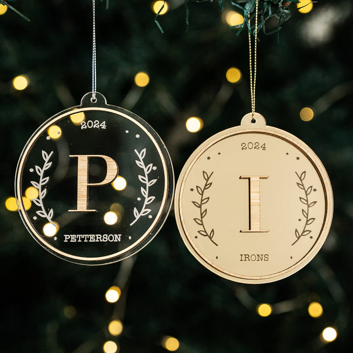 Two Monogram Christmas Baubles hang on the tree. Each bauble features a name, initial, 2024, and leafy borders—a personalised ornament and charming keepsake from The Monogram Christmas Bauble collection. Blurred lights glow softly behind.