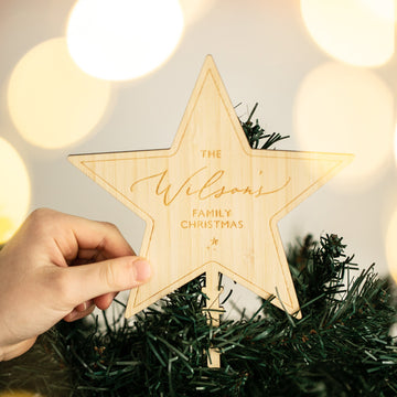 A hand places a Personalised Christmas Tree Topper reading "The Wilsons Family Christmas" on a green tree, with blurred soft yellow lights in the background.