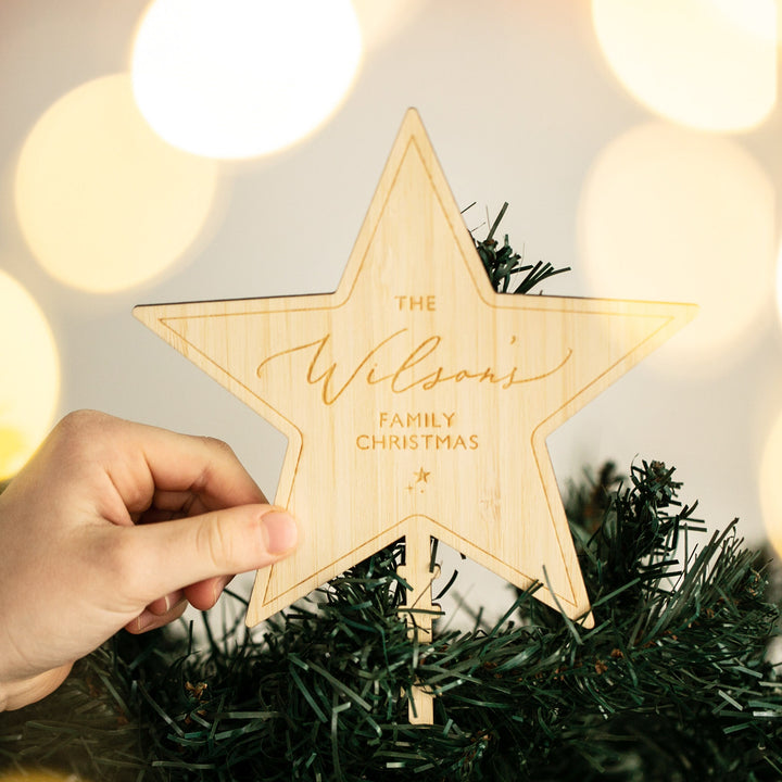 A hand places a Personalised Christmas Tree Topper reading "The Wilsons Family Christmas" on a green tree, with blurred soft yellow lights in the background.