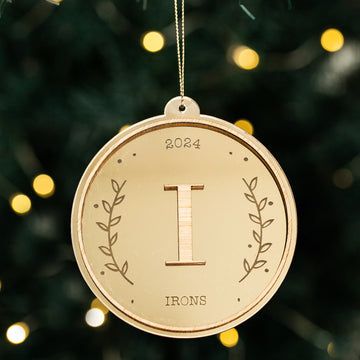 The Monogram Christmas Bauble is a round wooden ornament engraved with "2024," a large "I," "IRONS," and laurel branch designs—perfect as a unique personalised Christmas decoration.