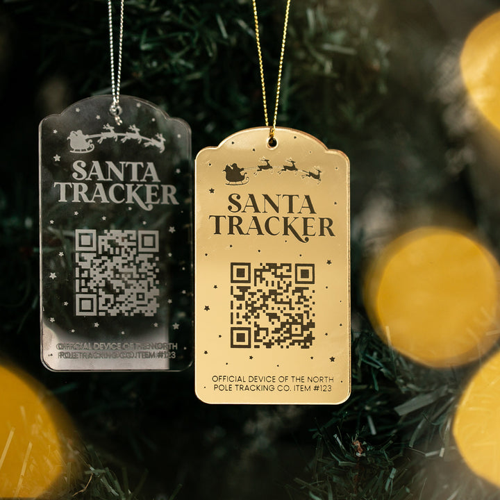 A clear and a gold hanging tag labeled Santa Tracker, each with a QR code, dangle before a Christmas tree’s blurred yellow lights—highlighting the interactive Santa Tracker Christmas Bauble ornament.