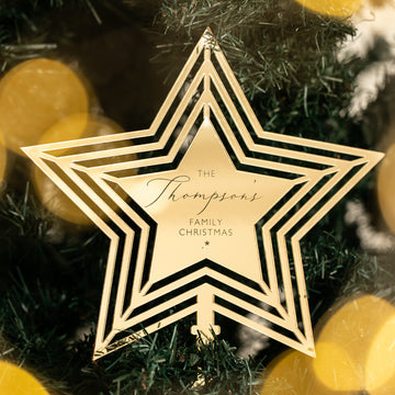 A Personalised Christmas Tree Topper engraved with "The Thompsons Family Christmas" hangs on a tree, surrounded by green branches and blurred yellow holiday lights.