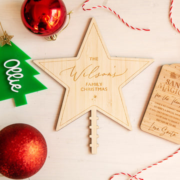 A Personalised Christmas Tree Topper, engraved with "The Wilsons Family Christmas," sits on a table among red ornaments, a green tree-shaped tag, and a wooden Santa Magic plaque.