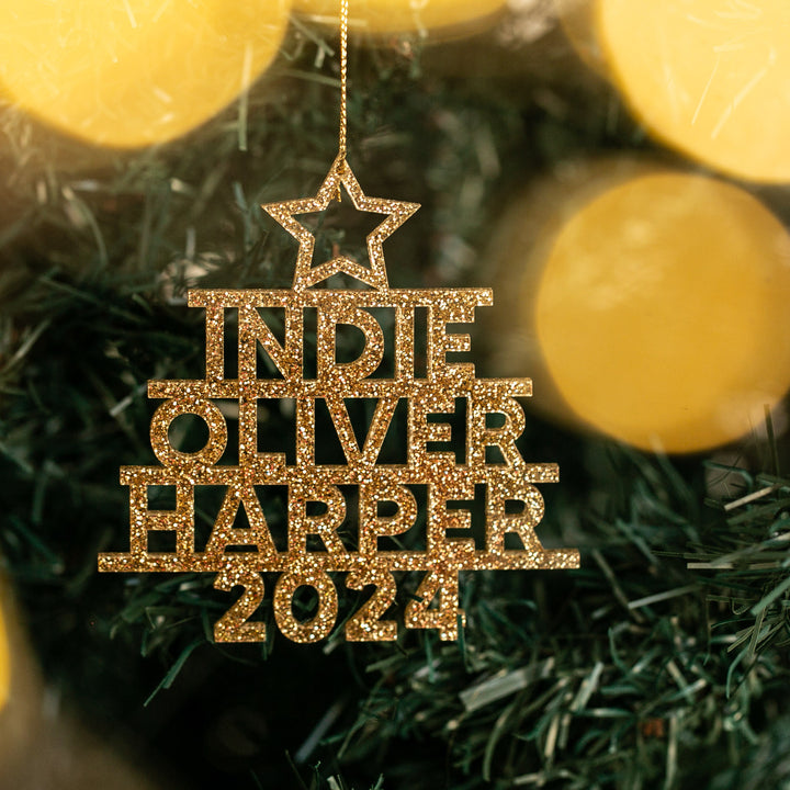 A gold glittery Whānau Christmas Tree Bauble, shaped like a tree with a star, hangs on a tree. The personalised ornament reads INDIE OLIVER HARPER 2024, set against softly glowing golden lights in the background.