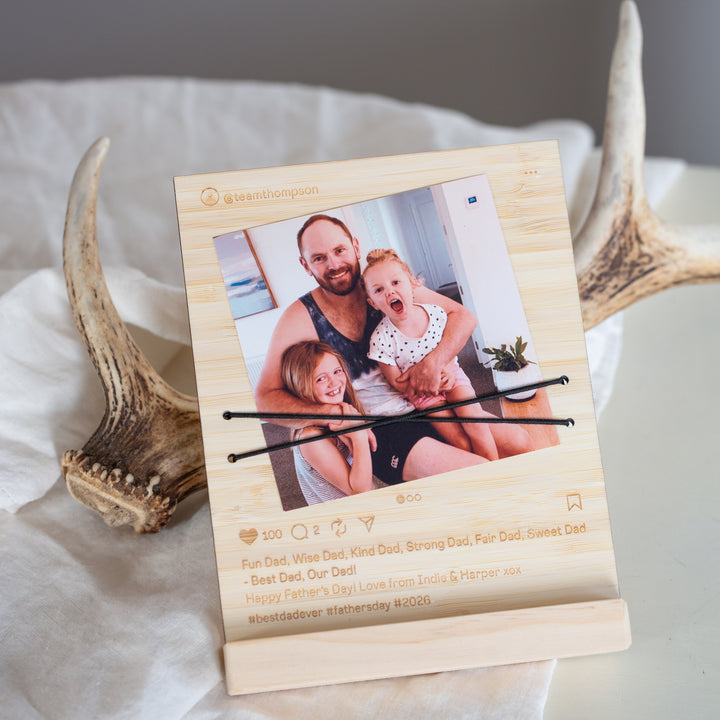 A personalized wooden photo frame styled on a white fabric surface with antlers in the background. The frame features a custom photo of a father and children, along with engraved text celebrating Father&