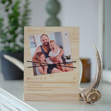 A personalized light wood photo frame with a printed social-media-style design, holding a family photo and two black elastic bands across the center. The frame is displayed on a white surface with a blurred indoor background and a decorative antler prop.