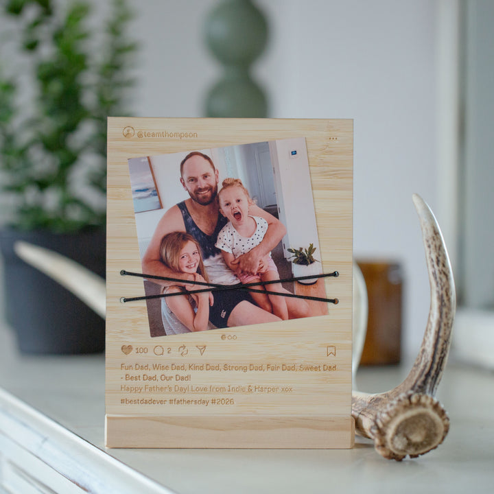 A personalized light wood photo frame with a printed social-media-style design, holding a family photo and two black elastic bands across the center. The frame is displayed on a white surface with a blurred indoor background and a decorative antler prop.
