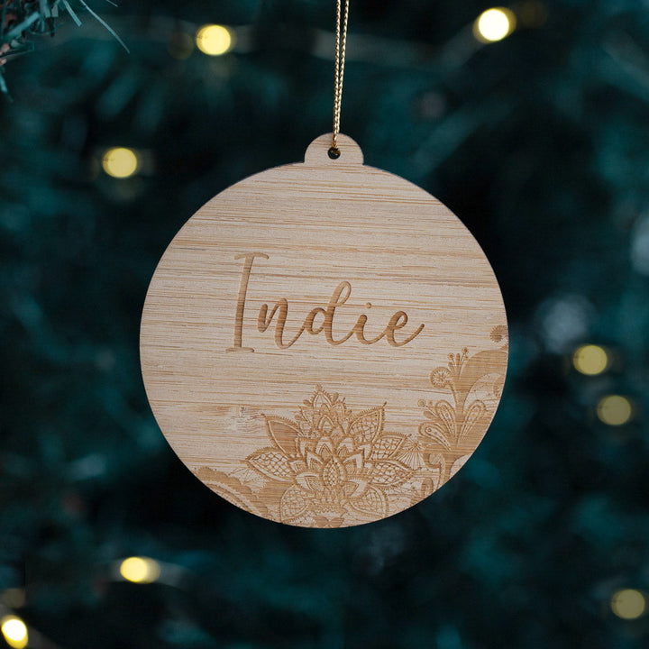 The Lace Christmas Bauble is a round, eco-friendly bamboo ornament on a gold string, engraved with "Indie" and a floral design, shown against blurred holiday lights and greenery in the background.