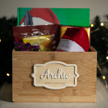 A Personalised Christmas Eve Box labeled "Archie Merry Christmas" sits on a table, brimming with festive treats, a Santa hat, and a green book. This special keepsake glows warmly amid twinkling lights and lush greenery in the background.