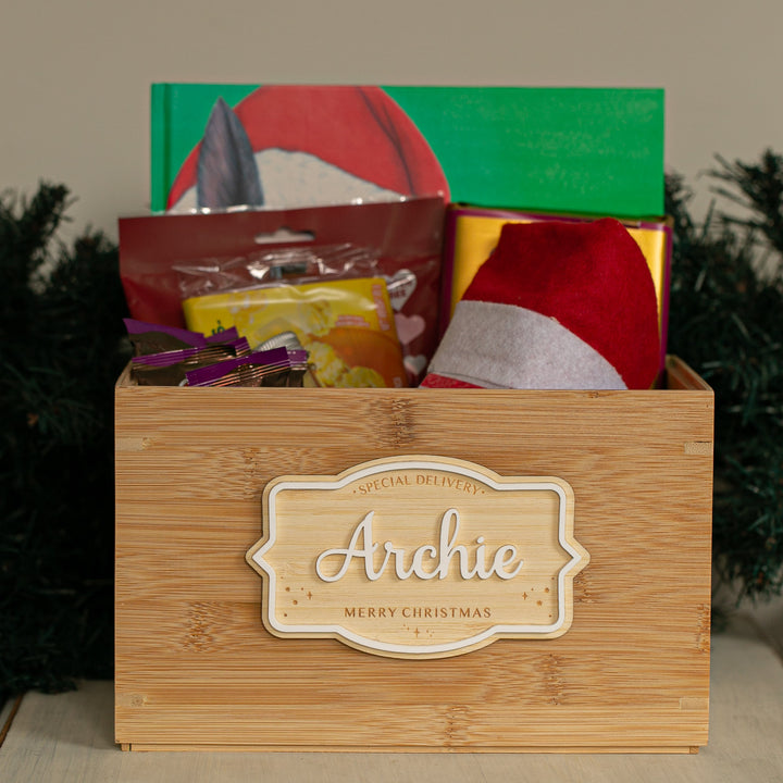 A Personalised Christmas Eve Box labeled "Archie Merry Christmas" is filled with festive items like a Santa hat, snacks, and a green book, making it a perfect holiday keepsake against greenery in the background.