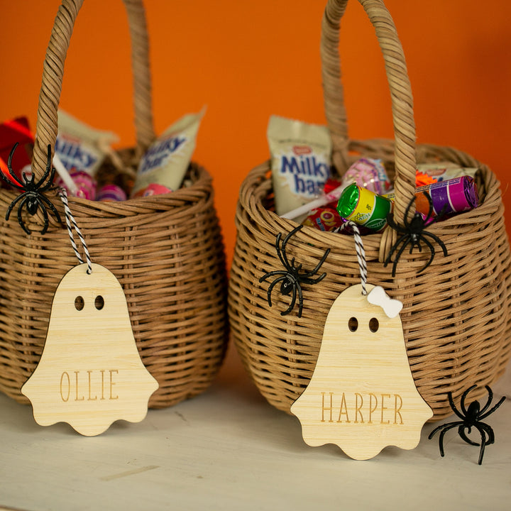 Two wicker baskets filled with Halloween candy, each featuring a Personalised Wooden Halloween Tag with names OLLIE and HARPER. Plastic spiders decorate the baskets, arranged on an orange background.