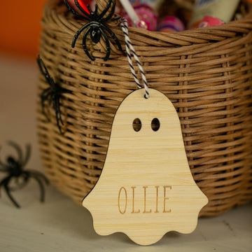 A Personalised Wooden Halloween Tag, engraved with "OLLIE," hangs from a wicker basket with black spiders and candy.