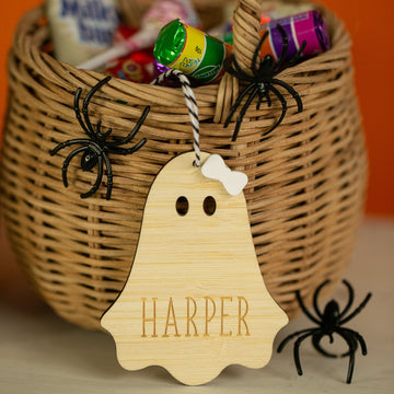 A wicker basket of Halloween candy with black plastic spiders is shown. In front, a Personalised Wooden Halloween Tag featuring the name HARPER hangs by a black-and-white string.