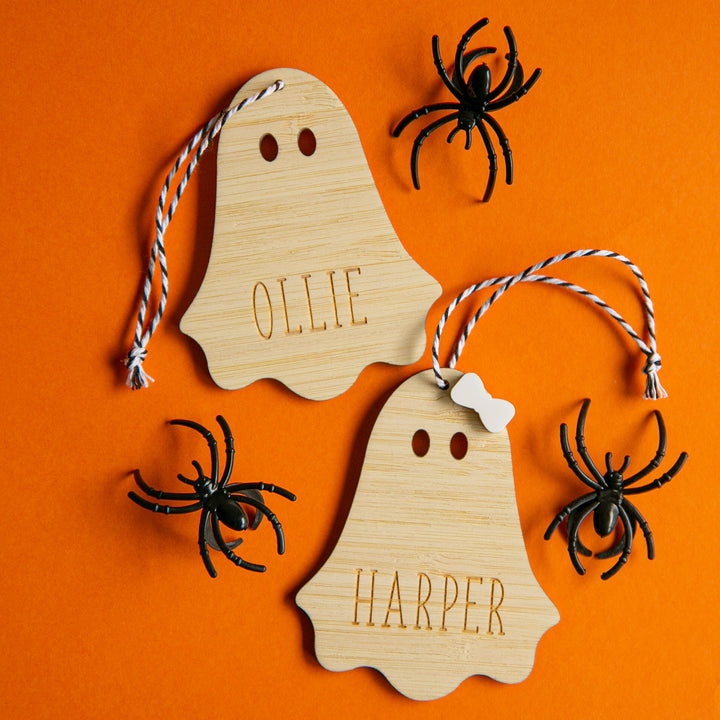 Two Personalised Wooden Halloween Tags, labelled OLLIE and HARPER, with string loops and one featuring a white bow, are displayed on an orange background surrounded by three black plastic spiders.