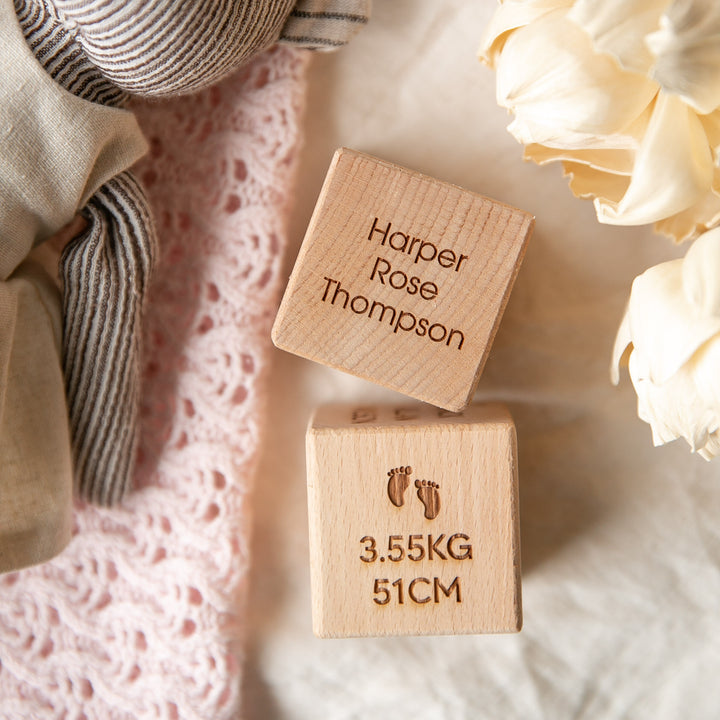 Two Personalised Wooden Blocks rest on a soft surface: one displays "Harper Rose Thompson," the other shows "3.55KG 51CM" with tiny footprints. A knit toy and cream flowers nearby make these blocks an ideal newborn gift.