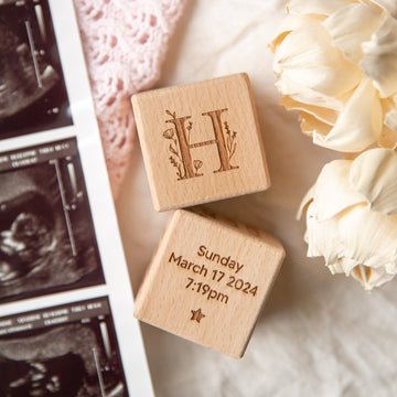 Two Personalised Wooden Blocks are shown: one engraved with a floral letter H as a baby keepsake, the other displaying "Sunday March 17 2024, 7:19pm." They’re beside dried flowers and ultrasound photos—a unique newborn gift.