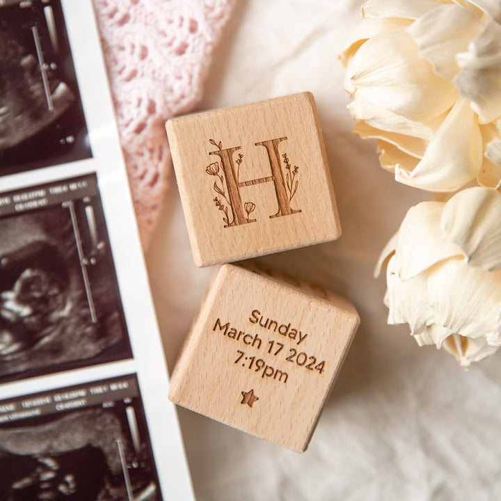 Two Personalised Wooden Blocks are shown: one engraved with a floral letter H as a baby keepsake, the other displaying "Sunday March 17 2024, 7:19pm." They’re beside dried flowers and ultrasound photos—a unique newborn gift.