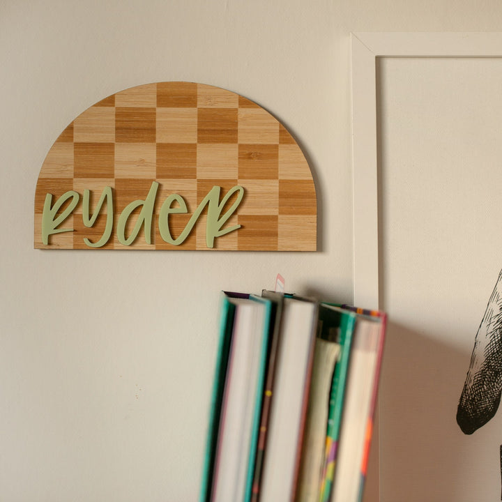A wooden half-circle Personalised Checkerboard Plaque with "ryder" in green script hangs above colorful books and a framed print, creating charming nursery decor.