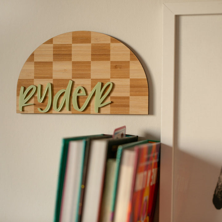 A Personalised Checkerboard Plaque with "ryder" in green cursive hangs above colorful books near a white door, bringing unique nursery decor to the space.