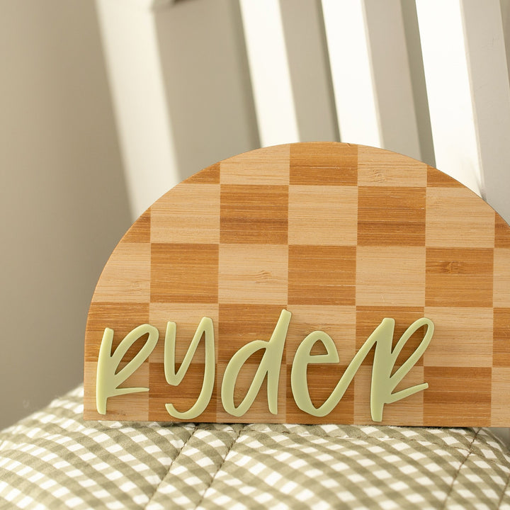 The Personalised Checkerboard Plaque features the name ryder in light green script on a wooden semicircle, shown leaning against a white chair with a green and white checkered cushion—ideal for unique nursery decor.