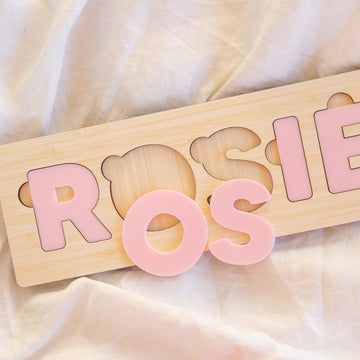 The Personalised Name Puzzles, made from eco-friendly bamboo, feature pale pink letters spelling ROSIE on a light fabric. The letter O is removed from this custom baby gift and positioned in front for playful interaction.