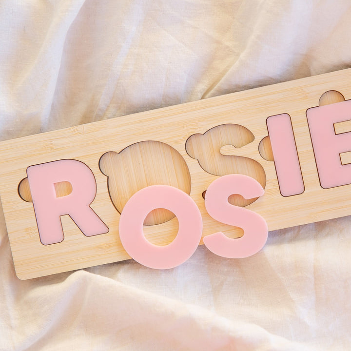 The Personalised Name Puzzles, made from eco-friendly bamboo, feature pale pink letters spelling ROSIE on a light fabric. The letter O is removed from this custom baby gift and positioned in front for playful interaction.