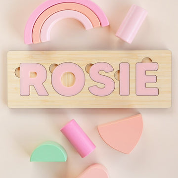 Personalised Name Puzzles: Spell ROSIE in pink letters with pastel geometric shapes and a rainbow on a light beige background. A custom baby gift crafted from eco-friendly bamboo.