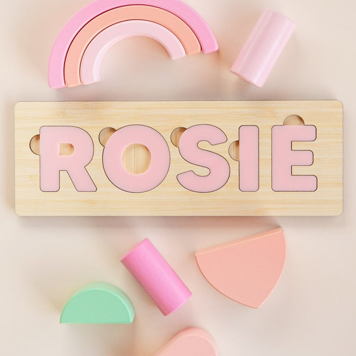 Personalised Name Puzzles: Spell ROSIE in pink letters with pastel geometric shapes and a rainbow on a light beige background. A custom baby gift crafted from eco-friendly bamboo.