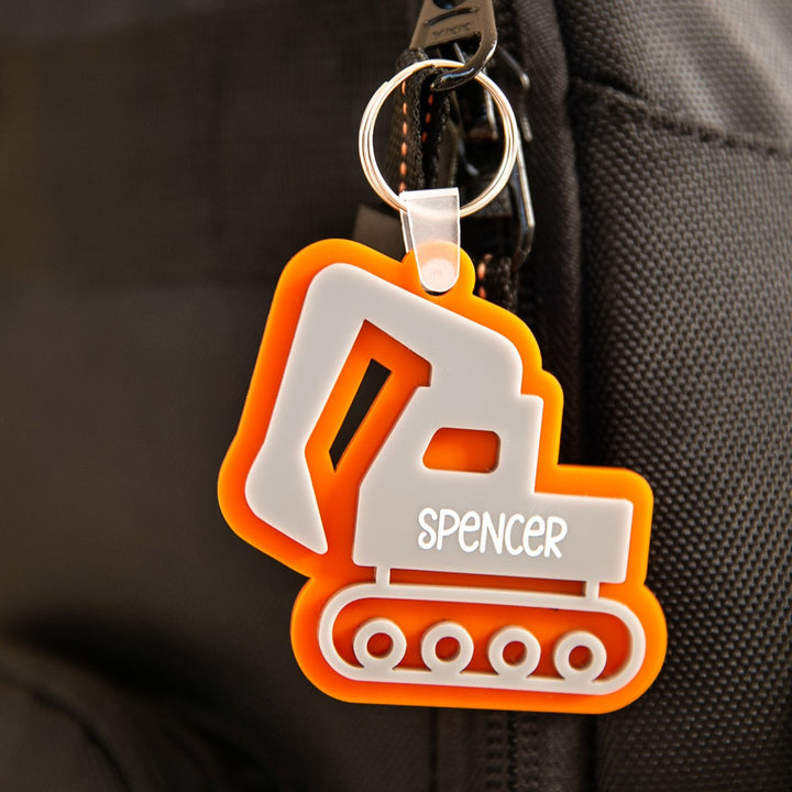 The Icon Personalised Bag Tag, shaped like an orange and white excavator with the name Spencer, is attached to a black backpack zipper—ideal as a kids’ bag tag.
