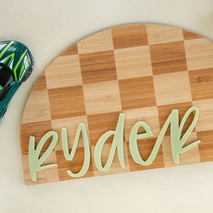 The Personalised Checkerboard Plaque is a wooden half-circle sign with a checkered pattern and the name Ryder in light green script, making it a charming nursery decor piece. A green toy car appears partially on the left in the image.