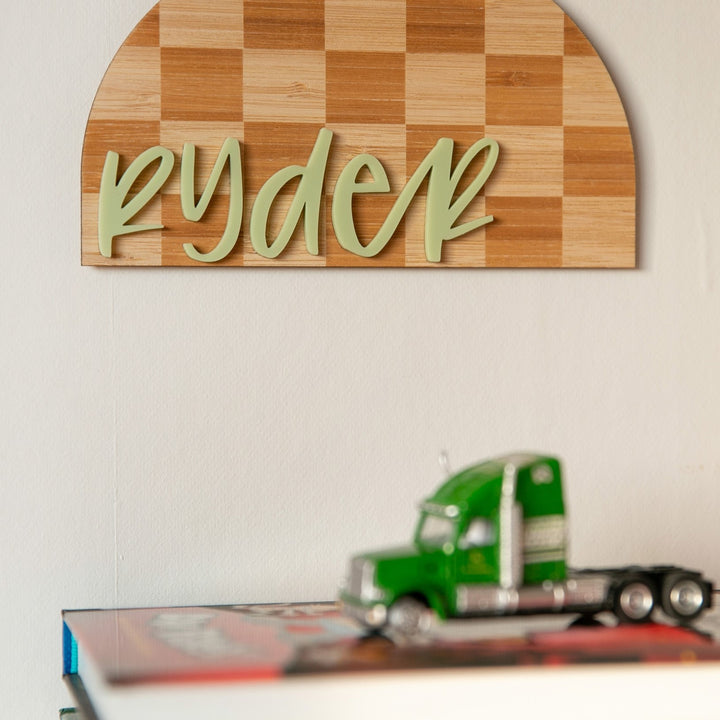 The Personalised Checkerboard Plaque features a custom name in green cursive above a green toy truck on stacked books—ideal nursery decor for adding a playful, personalised touch.