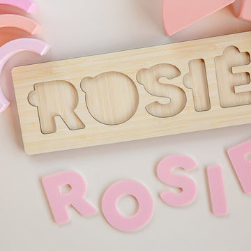 The Personalised Name Puzzles feature bear-themed, pink removable letters spelling a child’s name on a crisp white bamboo board. Eco-friendly and customisable, each puzzle includes curved pink pieces for a playful touch.