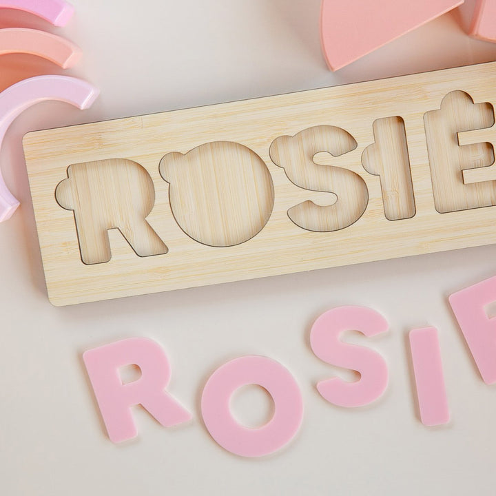 The Personalised Name Puzzles feature bear-themed, pink removable letters spelling a child’s name on a crisp white bamboo board. Eco-friendly and customisable, each puzzle includes curved pink pieces for a playful touch.