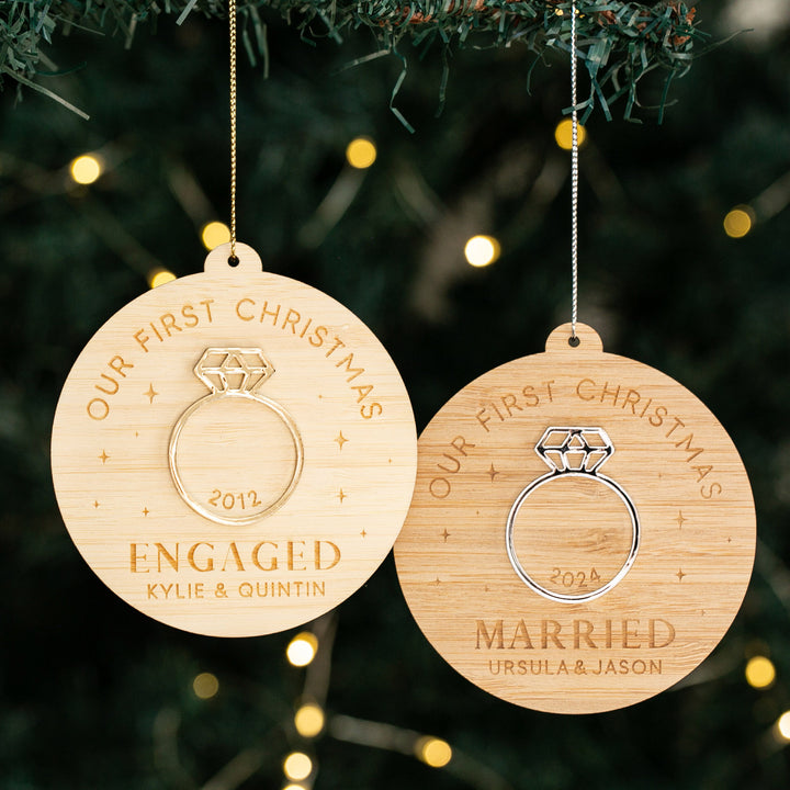 Two wooden Diamond Ring Christmas Bauble ornaments hang on a tree. One reads “Engaged 2022, Kylie & Quintin,” the other reads “Married 2024, Ursula & Jason.” Both feature “Our First Christmas” at the top—a personalised decoration.